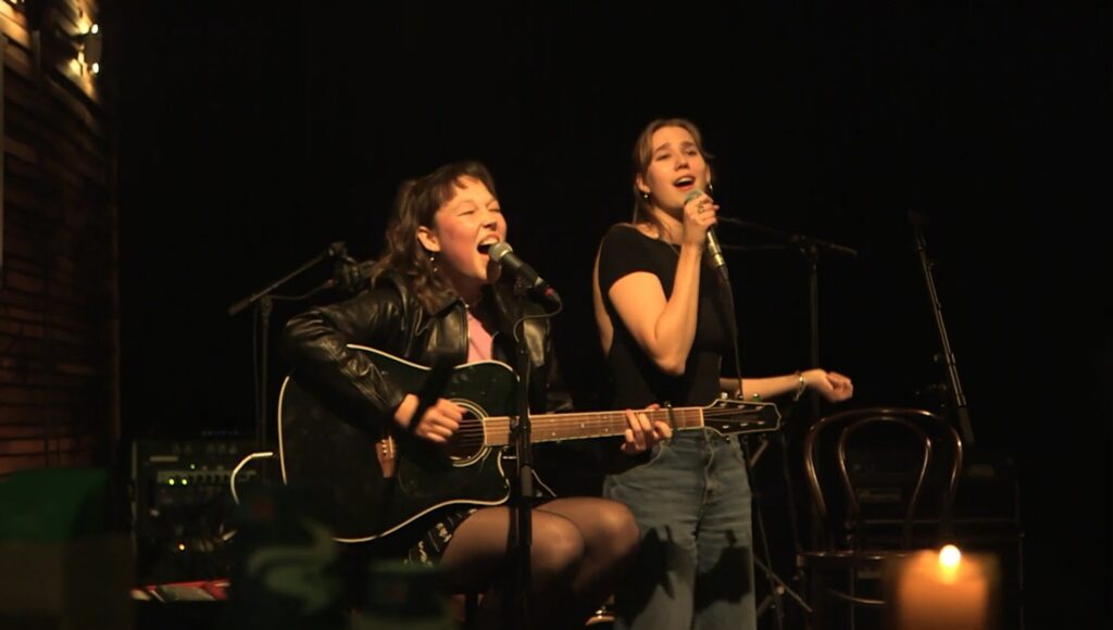 Twee zangeressen op het podium van het Cpunt Café, de ene speelt akoestische gitaar, de andere zingt in een microfoon